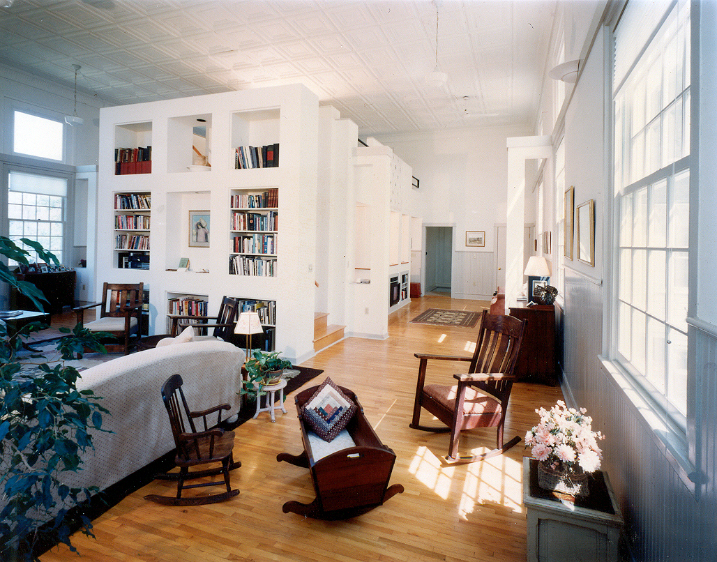 A grainy color photograph shows the interior of a church house that has been converted to a private residence. The view is from the first level looking toward the back of the house. There is a central enclosed kitchen with stairs leading up to a second level open studio. 