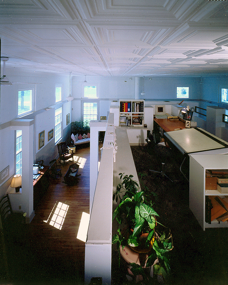 A grainy color photograph showing the interior of a church house that has been converted to a private residence. The view is from the second level and shows one large open space that has been organized by the construction of a central enclosed kitchen on the first level and study on the second level. 